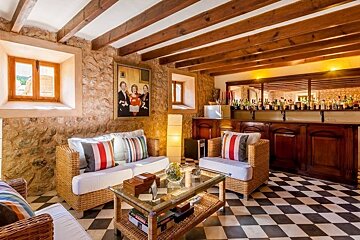 A rustic living room with stone walls, wooden beams, wicker furniture, checkered floor, and a well-stocked bar.