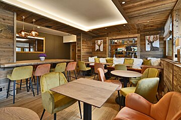 A cozy, modern rustic lounge featuring warm wood-paneled walls, a bar, and inviting seating with colorful velvet chairs in shades of yellow and orange.
