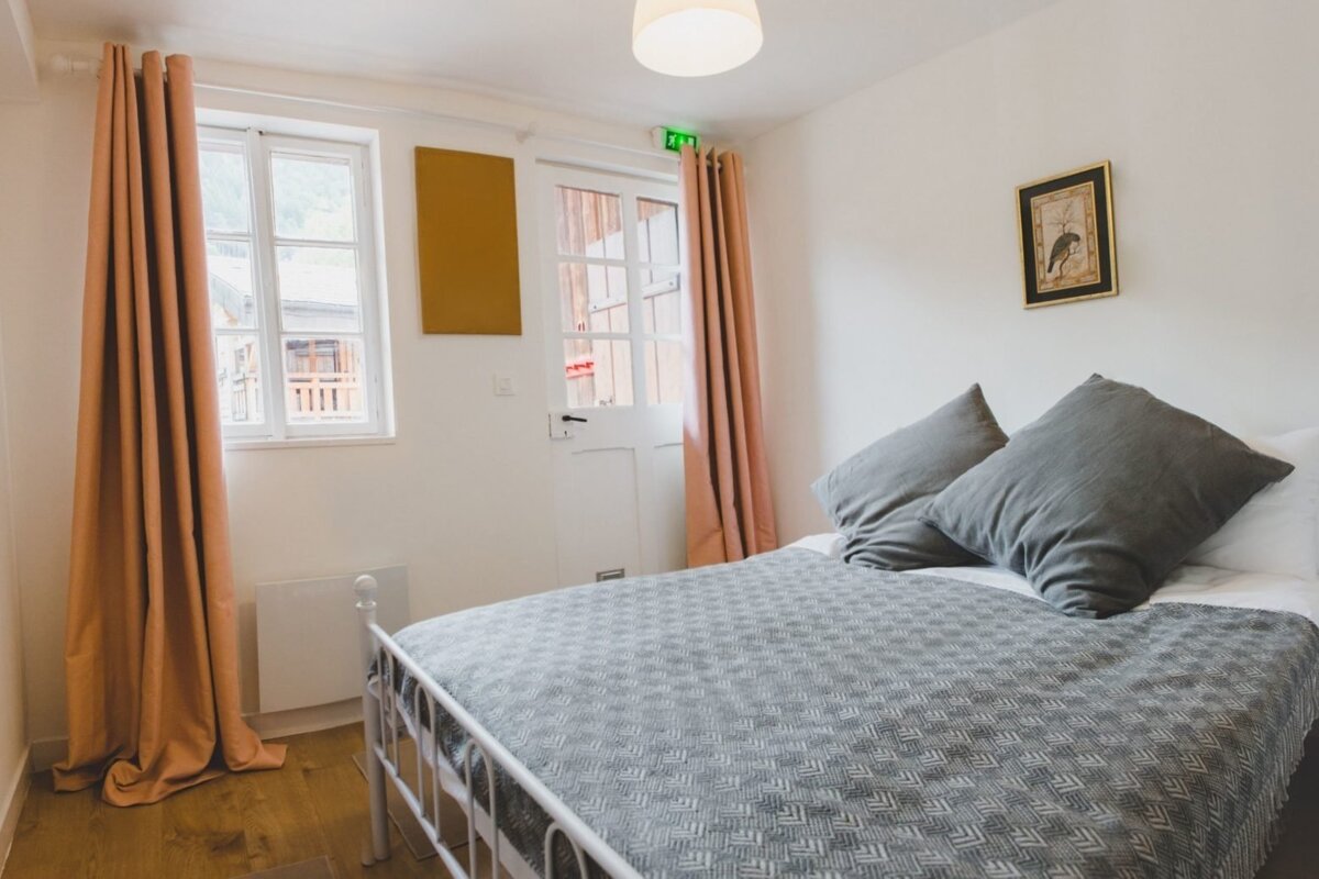 Cozy bedroom with white walls, a white bed frame, grey bedding, and peach curtains. A window and door offer natural light and exterior views.