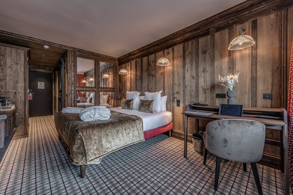A cozy hotel room with wood-paneled walls, a large bed with a white bathrobe, and a plaid carpet. A desk with a tablet is visible.