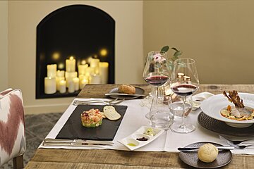A romantic dinner table set with appetizers, a main course, red wine, and a cozy fireplace full of glowing candles.