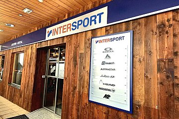 An Intersport store entrance with a rustic wooden facade. Signs advertise rentals and feature popular sports brands such as Oakley, Head, and Rossignol.