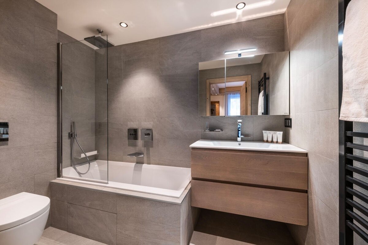 Stylish gray-tiled bathroom featuring a tub/shower combo, modern wooden vanity with sink and mirror, and a sleek toilet.