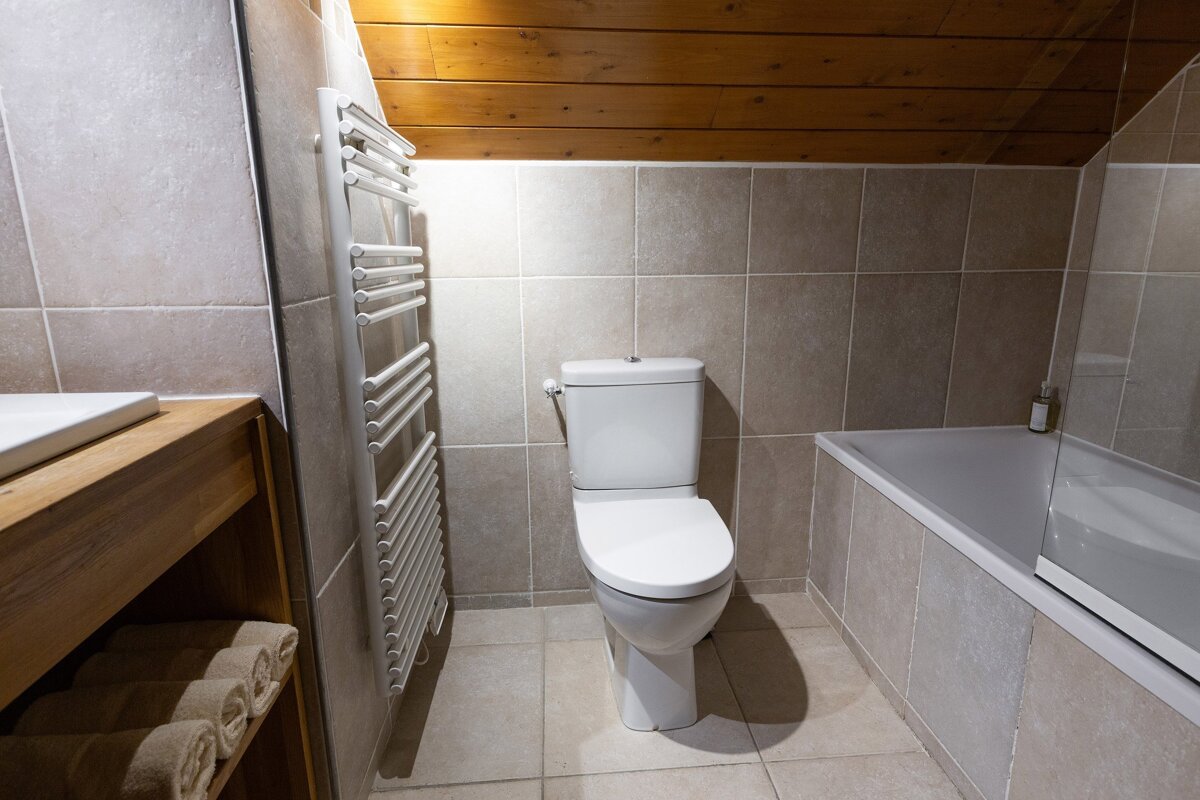 A modern tiled bathroom with a toilet, bathtub, and towel warmer under a warm wooden sloped ceiling. A sink vanity is partially visible.