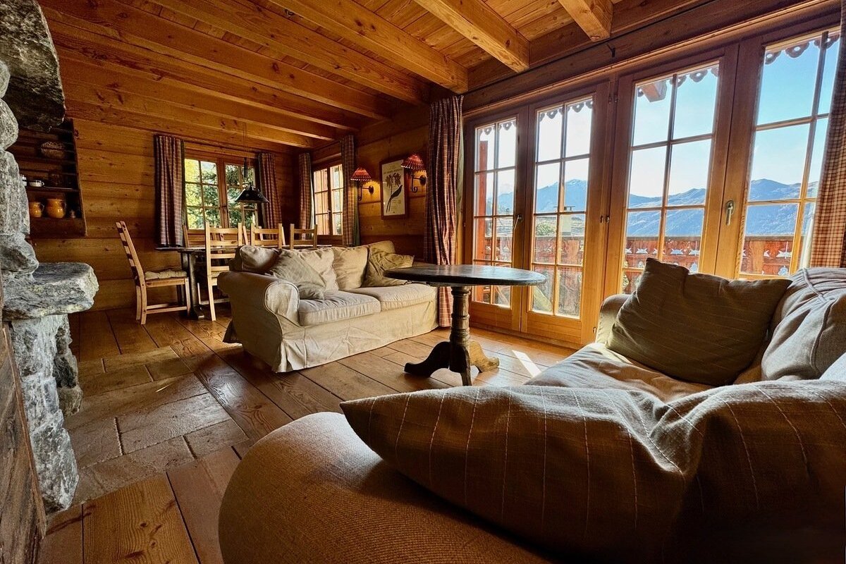 Warm, wood-paneled chalet living room with cozy sofas, a dining area, and large windows revealing stunning mountain vistas.
