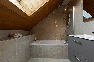 A modern attic bathroom featuring a tiled bathtub with a shower screen, a vanity, and a sloped wooden ceiling with a skylight. Towels sit on a shelf.