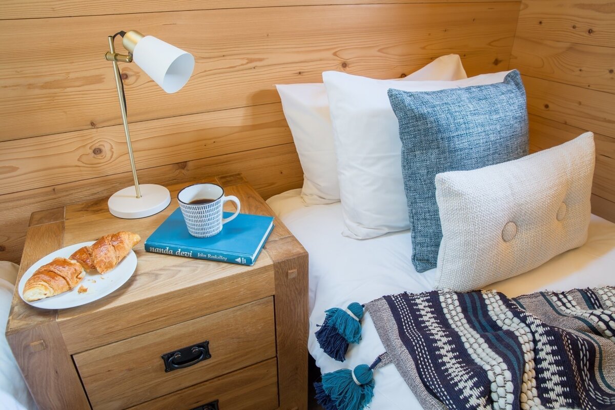 Cozy bedside table with croissants, coffee, a book, and a lamp beside a bed with pillows and a tasseled blanket against a wooden wall.