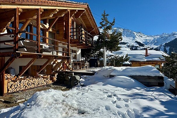 A charming wooden chalet with stacked firewood, covered in snow on a sunny day, against a backdrop of snow-capped mountains.