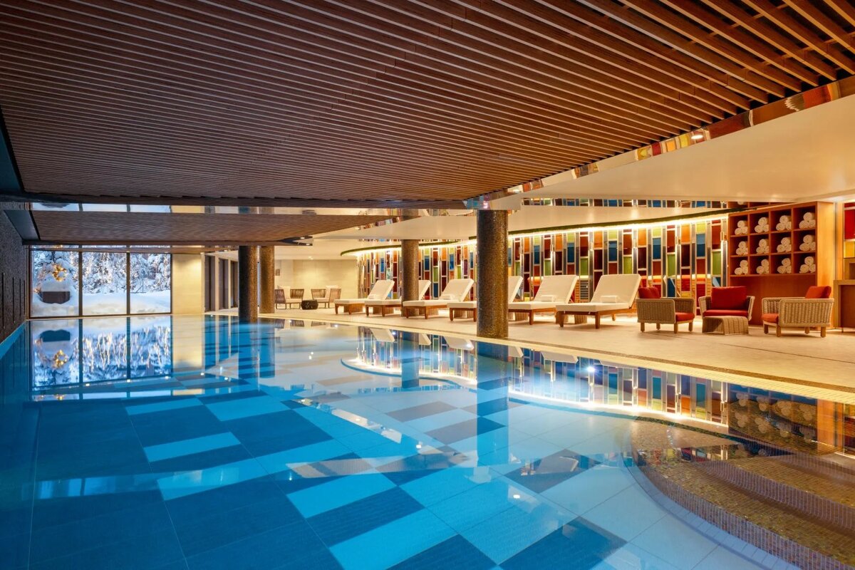 A luxurious indoor pool with a vibrant tiled wall, wooden ceiling, lounge chairs, and a window offering a view of a snowy landscape outside.