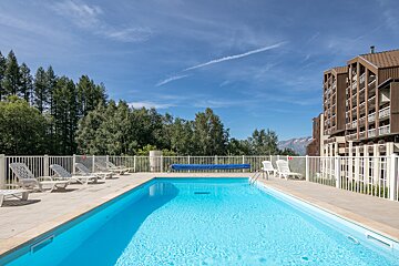 A bright blue swimming pool with lounge chairs and a white fence, surrounded by green trees and a large building under a clear sky with distant mountains.