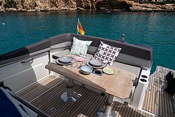 Boat's aft deck with a laid table, cushions, and German flag, overlooking serene turquoise waters, a rocky coastline, and a distant paddleboarder.