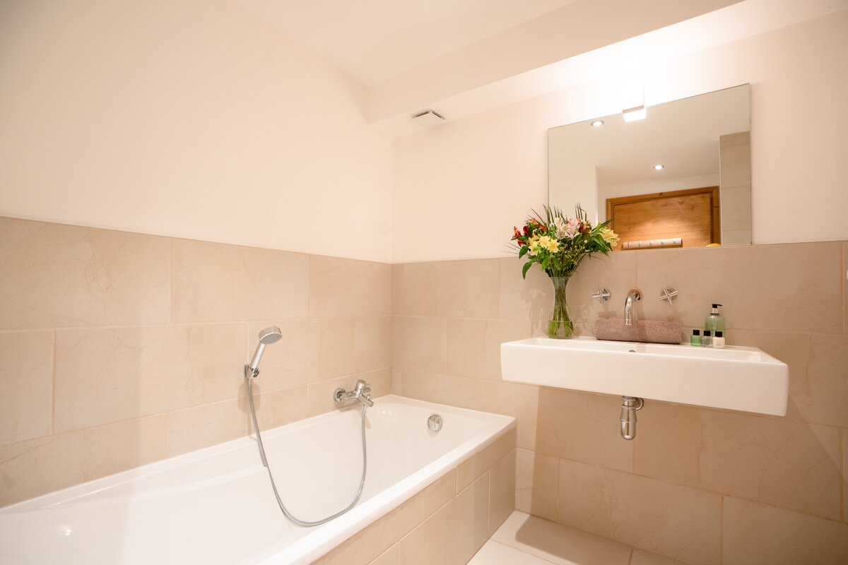 A modern, tiled bathroom with a white bathtub and a floating sink. A vase of colorful flowers sits by the sink, below a large mirror.