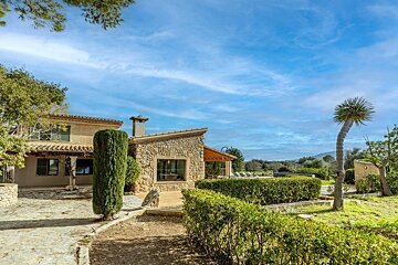 A rustic stone villa with a paved patio, lush hedges, and a tall palm tree, set against a bright blue sky and green hills.