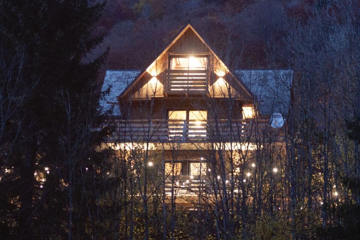 A cozy, brightly lit wooden cabin with balconies, nestled in a forest of bare trees with sparkling string lights at twilight.