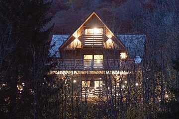 A cozy, brightly lit wooden cabin with balconies, nestled in a forest of bare trees with sparkling string lights at twilight.