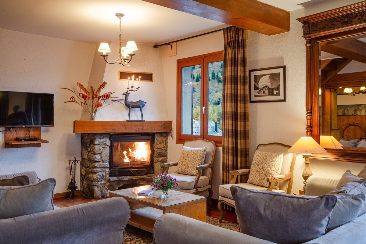 A cozy living room features a lit stone fireplace, comfortable sofas and armchairs, a TV, and a window with a mountain view. Warm lighting adds to the inviting atmosphere.