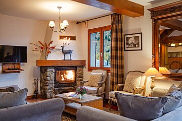 A cozy living room features a lit stone fireplace, comfortable sofas and armchairs, a TV, and a window with a mountain view. Warm lighting adds to the inviting atmosphere.
