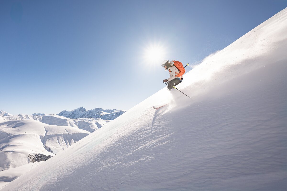 Three people in ski gear embrace on a snowy mountain, admiring a sunset over peaks and a circular lodge below.