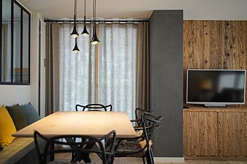 A modern dining area with a light wooden table, black chairs, and contemporary pendant lights. Rustic wood paneling and a TV complement beige curtains.