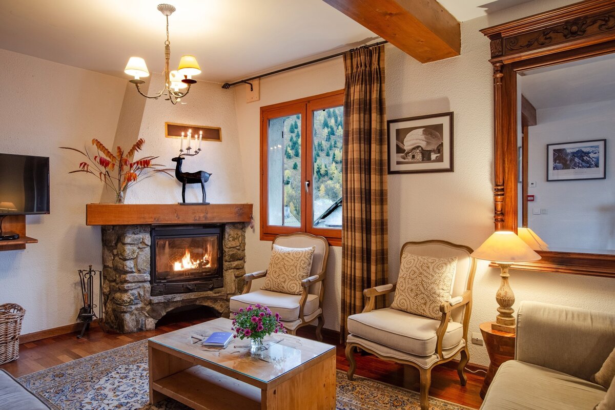 A warm, rustic living room with a lit stone fireplace, wooden beams, plush armchairs, and a window revealing a mountain view, creating a cozy atmosphere.