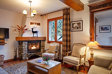 A warm, rustic living room with a lit stone fireplace, wooden beams, plush armchairs, and a window revealing a mountain view, creating a cozy atmosphere.