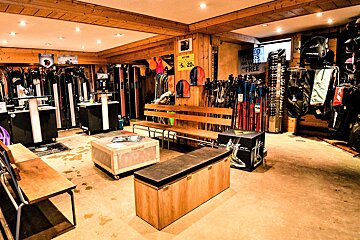 An indoor ski and snowboard shop, featuring racks of skis, poles, and gear. Wooden ceiling beams, walls, and benches create a rustic ambiance with warm lighting.