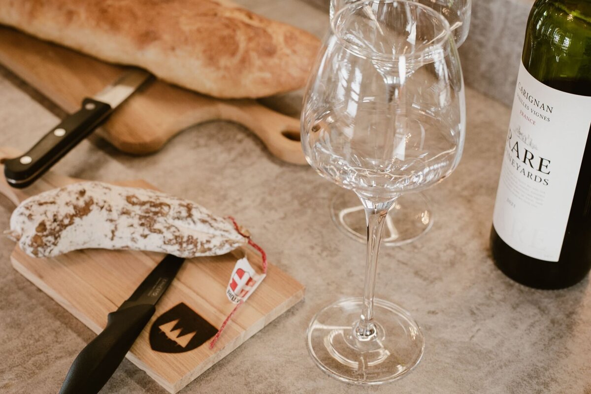 A rustic spread with baguette, salami, a knife on cutting boards, two empty wine glasses, and a bottle of red wine, ready for a cozy meal.