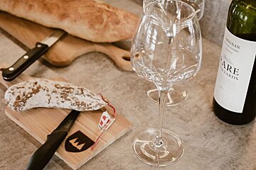 A rustic spread with baguette, salami, a knife on cutting boards, two empty wine glasses, and a bottle of red wine, ready for a cozy meal.