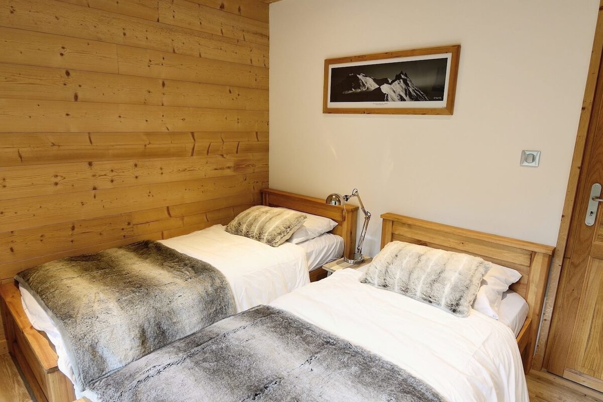 A cozy bedroom features two twin beds with wooden frames and faux fur bedding. A wooden paneled wall complements a white wall with a mountain photo.