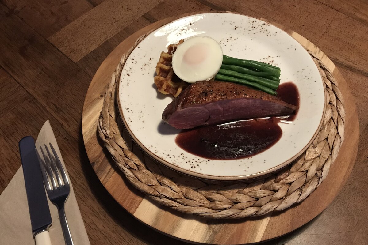 Pan-seared duck breast with a dark sauce, waffle, poached egg, and green beans on a plate. Served on a woven placemat with cutlery.