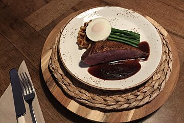 Pan-seared duck breast with a dark sauce, waffle, poached egg, and green beans on a plate. Served on a woven placemat with cutlery.