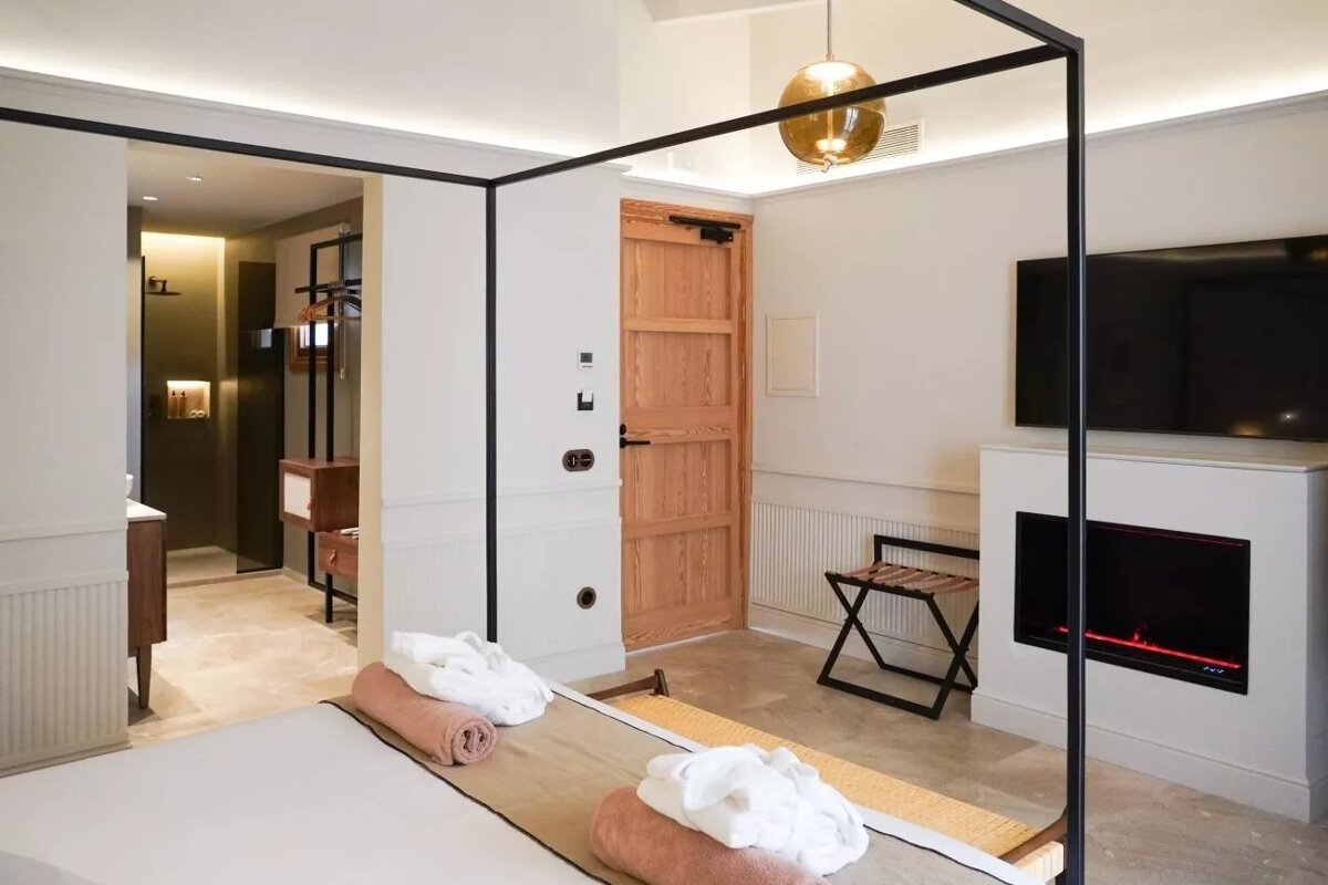 A modern hotel room with a black four-poster bed frame, white walls, a TV over a fireplace, and an adjoining bathroom/dressing area.