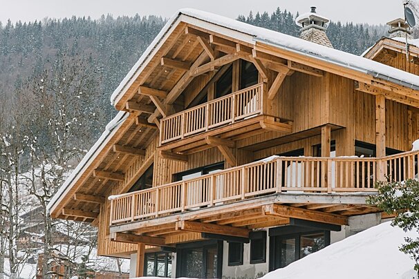 A large, modern wooden chalet with snow-covered roofs and multiple balconies, set against a backdrop of a snowy mountain forest.