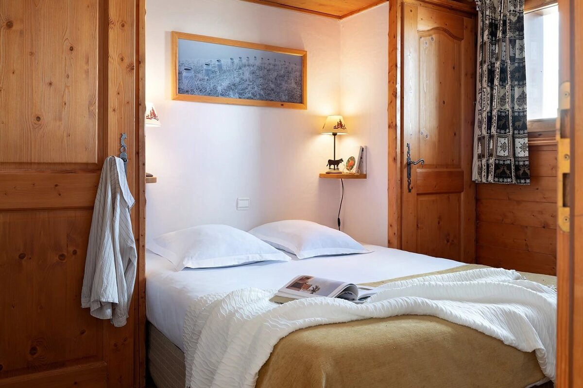 A cozy, rustic bedroom with wooden walls and doors, featuring a double bed with white linens, a bedside lamp, and a framed picture.