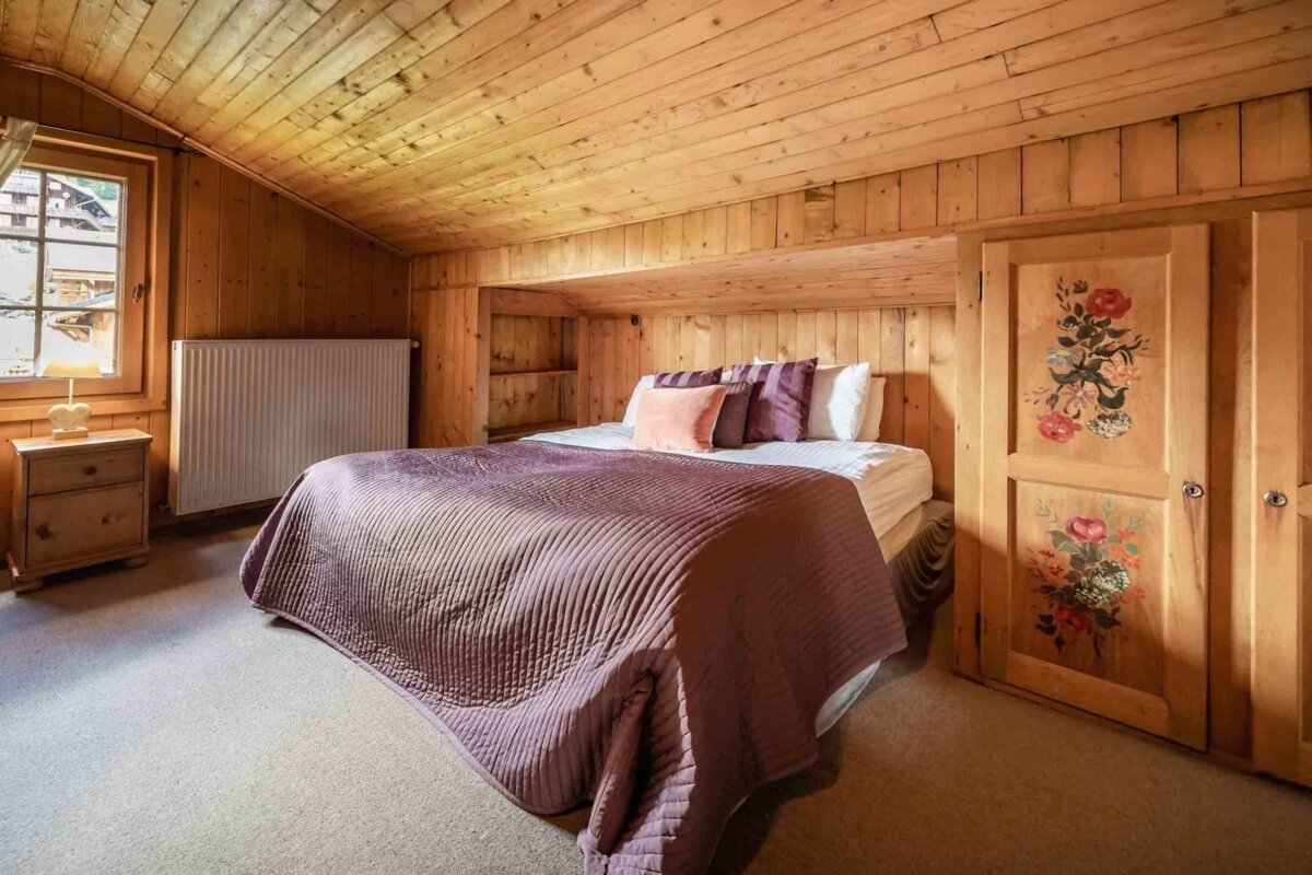 Cozy wood-paneled bedroom with a bed, purple bedding, a window, and a unique cabinet adorned with floral paintings.