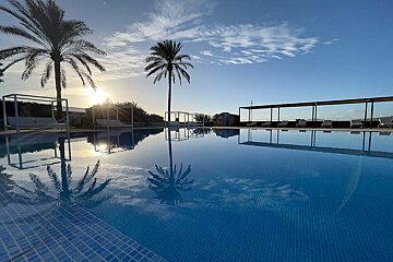 A serene blue-tiled pool reflects palm trees and the radiant sun at sunrise/sunset, under a clear sky. Lounge beds are visible nearby.