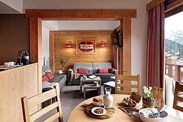A warm, wood-paneled chalet apartment with a cozy living area, dining table with breakfast items, and a balcony overlooking a snowy mountain village.