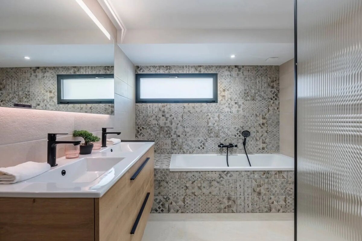 Modern bathroom with a light wood double vanity, black faucets, and a bathtub set against patterned tiles.