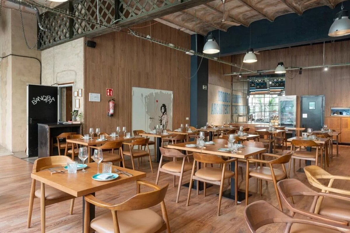 An industrial-chic restaurant with numerous wooden tables and chairs, set for dining. Features high ceilings, exposed elements, and modern decor.