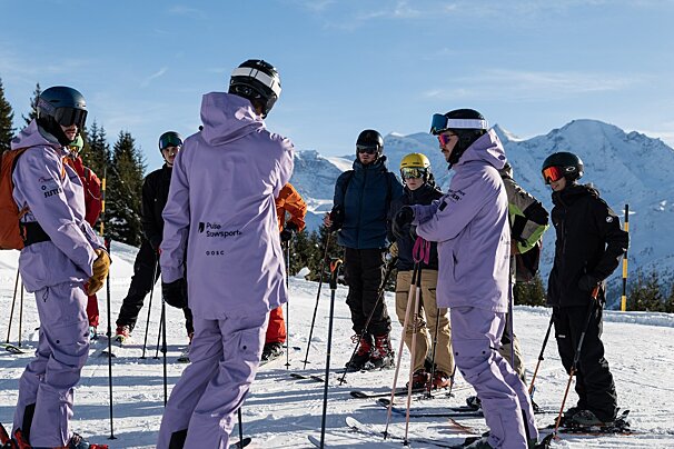 A group of skiers wearing helmets and goggles, including several in purple suits, stand on a sunny, snow-covered mountain with distant peaks.
