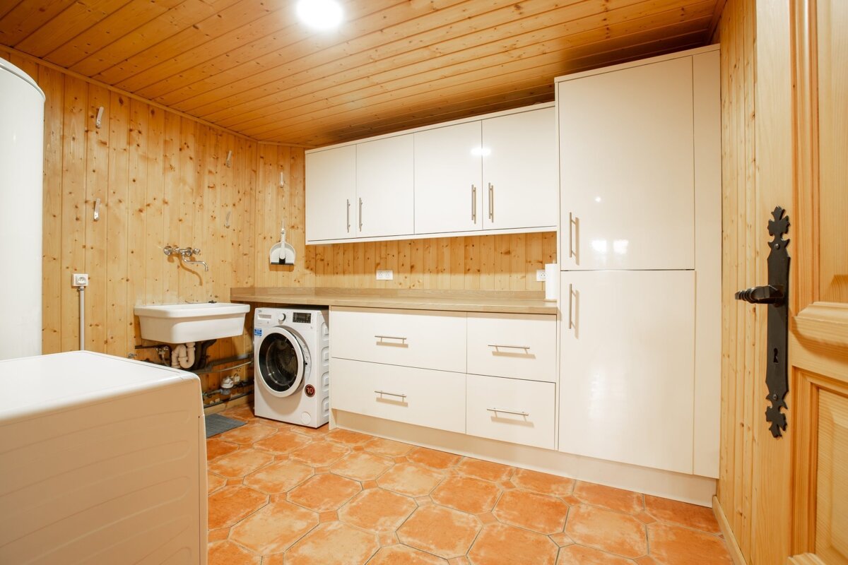 A wood-paneled utility room with white cabinets, a washing machine, a sink, a large water heater, and warm terracotta tile flooring.