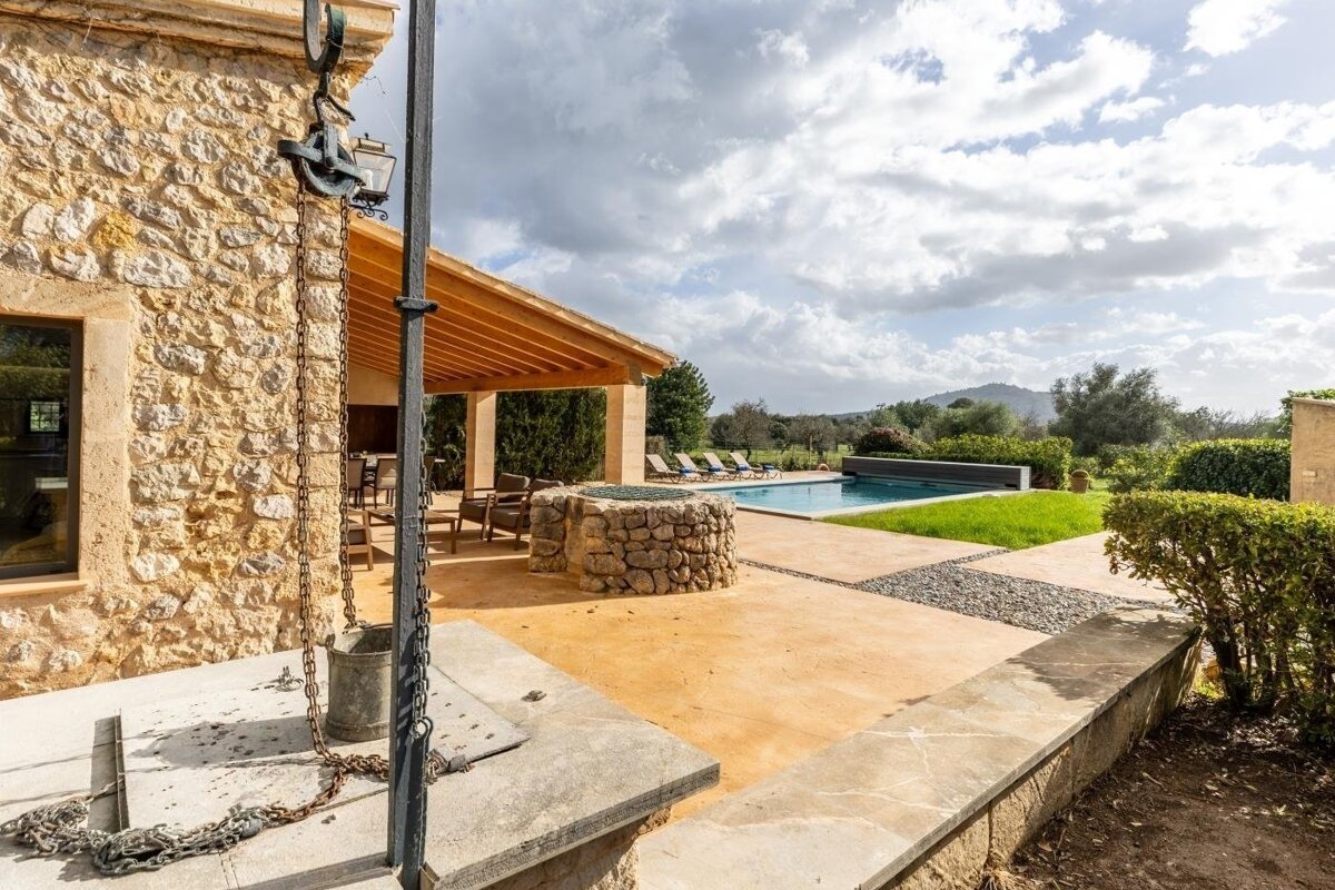 A rustic stone house with an old well, covered patio, and a swimming pool, all set within a green garden under a cloudy sky.