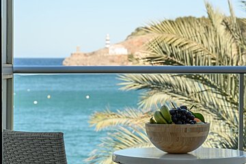 Balcony view of a vivid blue sea, a lighthouse on a distant hill, and palm trees. A bowl of fresh fruit sits on a table beside a wicker chair.