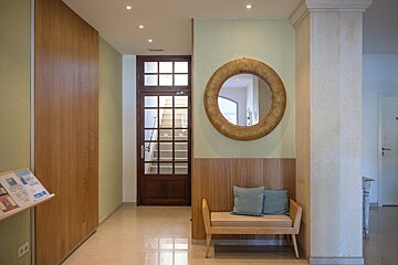 A welcoming hallway with light green walls, wood paneling, a round woven mirror, and a wooden bench with blue cushions. A glass-paned door leads to stairs.