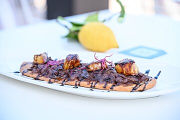 A savory flatbread on a white plate, topped with rich caramelized onions, three seared scallops, and red microgreens. A blurred lemon with leaves behind.