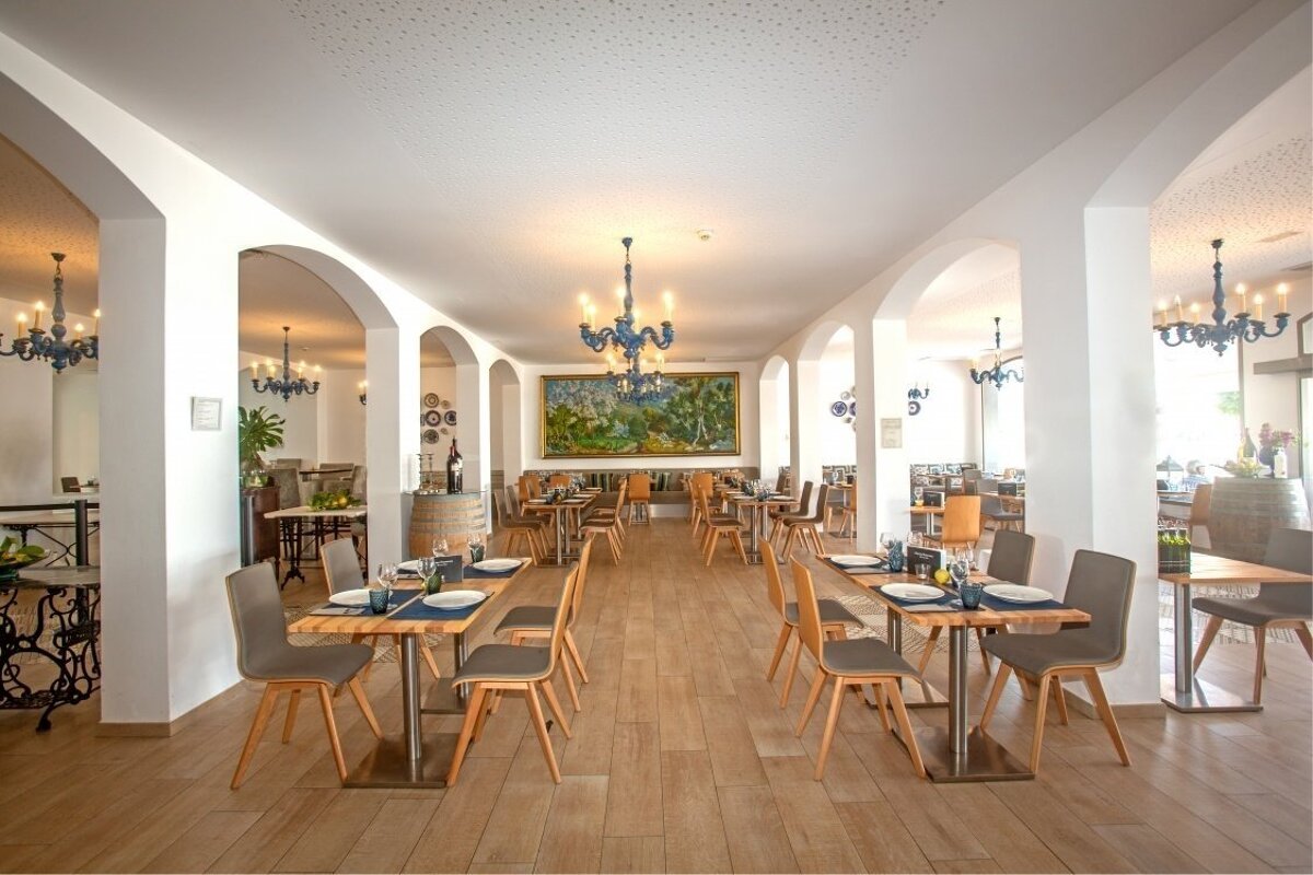 Spacious, elegant restaurant interior with arches, wooden tables and chairs, blue chandeliers, and a large landscape painting on white walls.