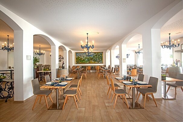 Spacious, elegant restaurant interior with arches, wooden tables and chairs, blue chandeliers, and a large landscape painting on white walls.