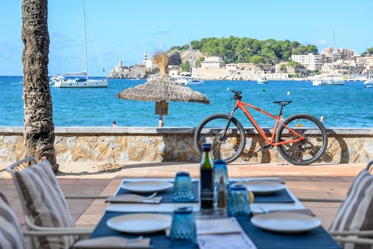Restaurant table by the sea, an orange bike, boats, and a lighthouse under a clear blue sky.