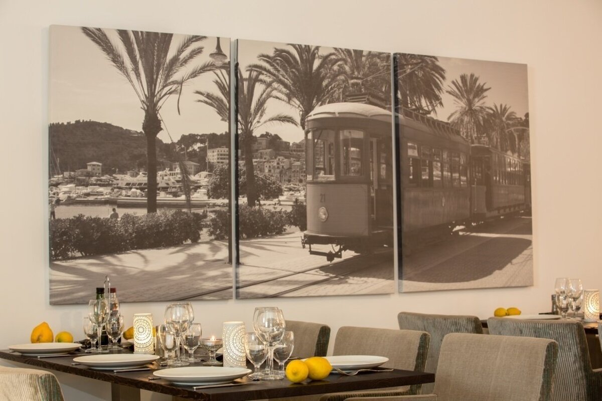 A restaurant dining area features a table set with glasses and lemons, beneath a large sepia-toned triptych art of a tram, palm trees, and a harbor.