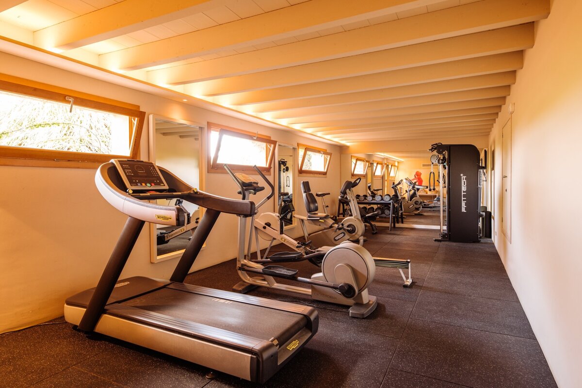 A brightly lit, spacious gym with various exercise machines including a treadmill, elliptical, and weights, under a ceiling with exposed wooden beams.
